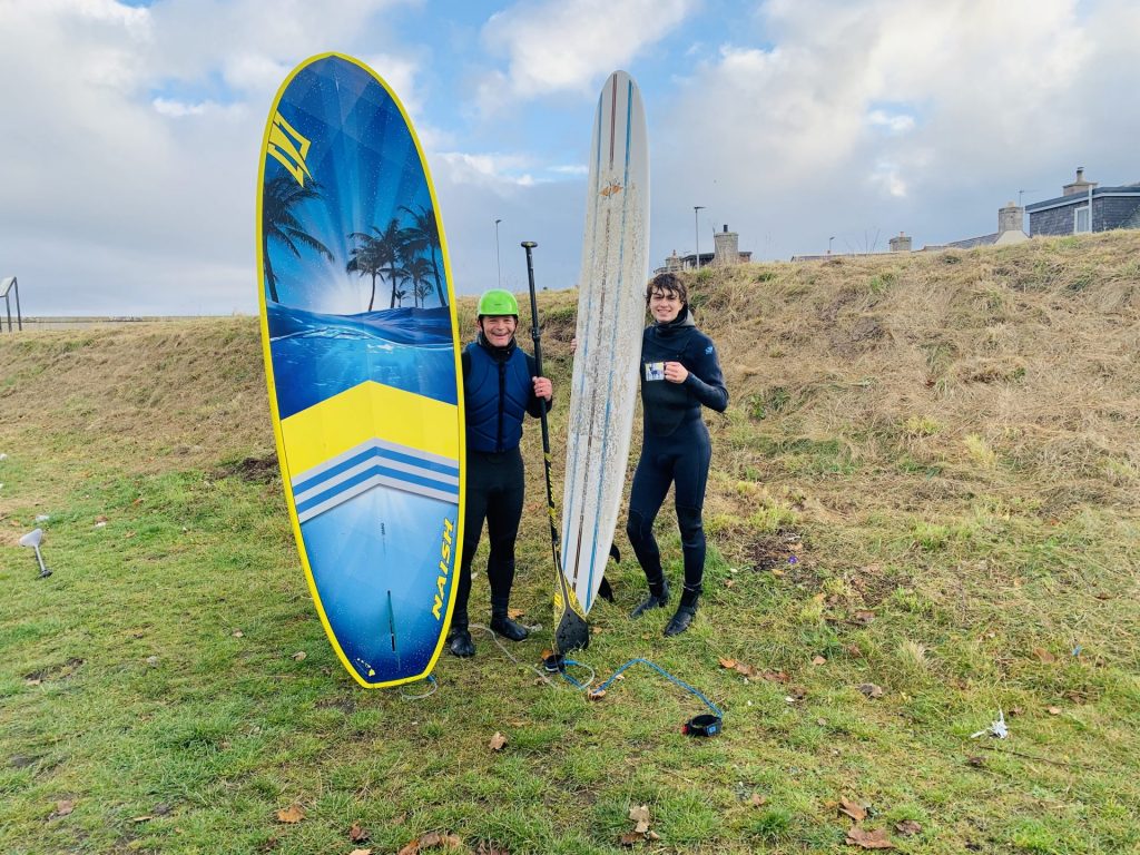 Two people hold surfboards upright, standing on grass near the seacoast. 