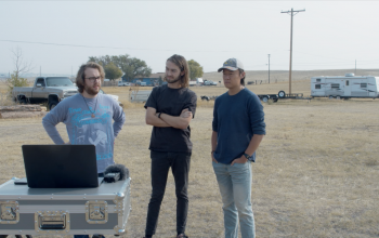 Sam, Nicholas, and Eric stand outside behind a laptop with a field behind them.