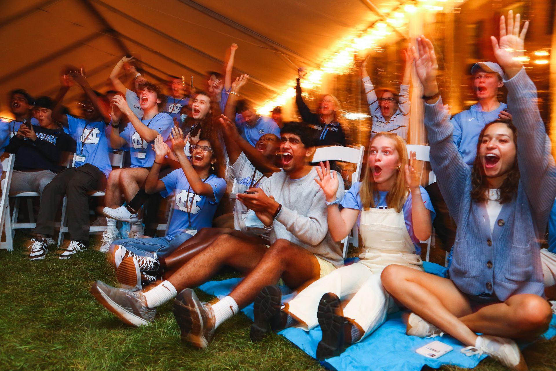 A group of students sitting on the grass cheer on the Tar Heels on television.