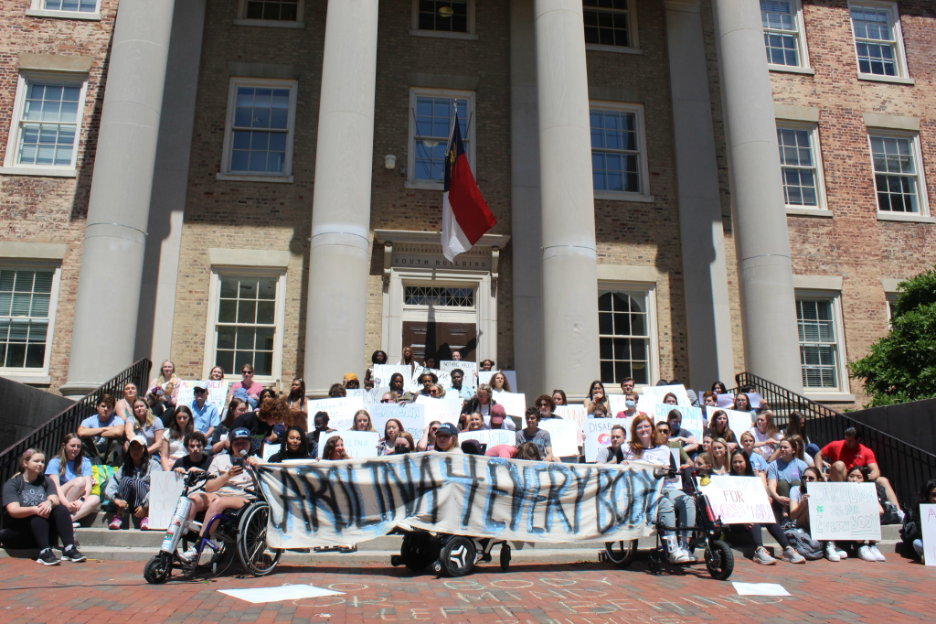 A group of students sit on the steps of South Building on a sunny day as part of a protest. There is a North Carolina flag on the door. Three students in wheelchairs hold a “CAROLINA 4 EVERYBODY” banner in the front of the group.