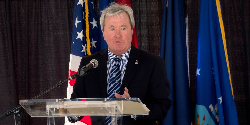 Haywood speaks behind a podium with a mic. There are patriotic flags behind him. He is wearing a striped shirt and tie, and a navy blazer.
