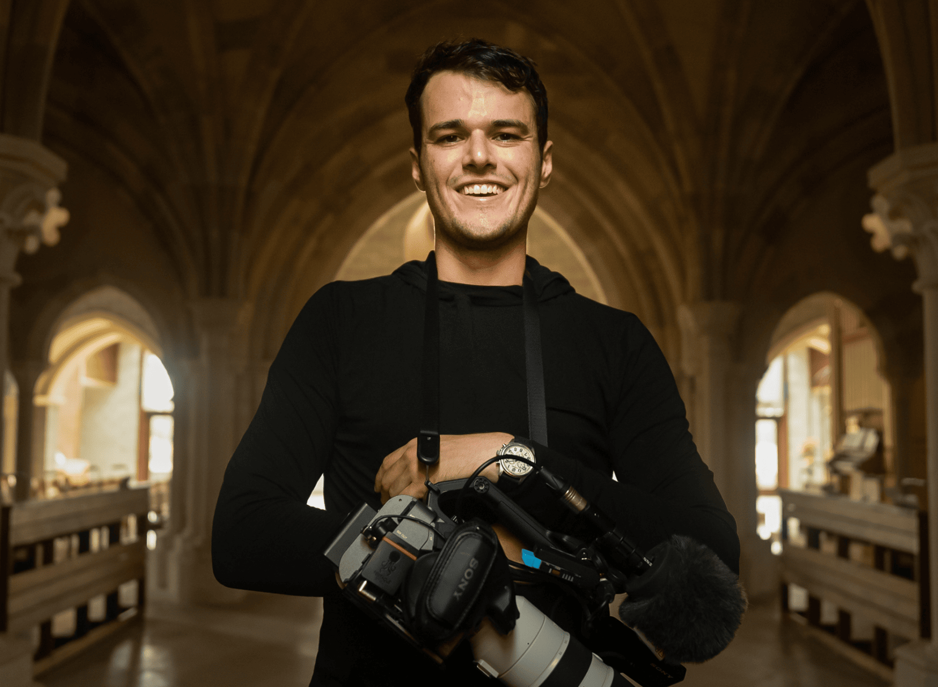 Rob Gourley ’18 smiles wearing a black long-sleeved shirt. His camera equipment is hanging around his neck.