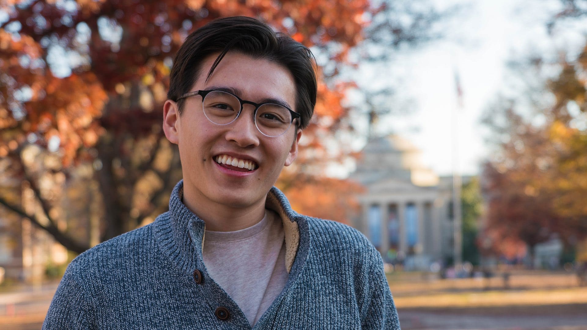 Sean Nguyen ’21 smile for the camera on Carolina's campus during the fall. There are trees and Graham Memorial in the background. Sean is wearing a grey sweater.
