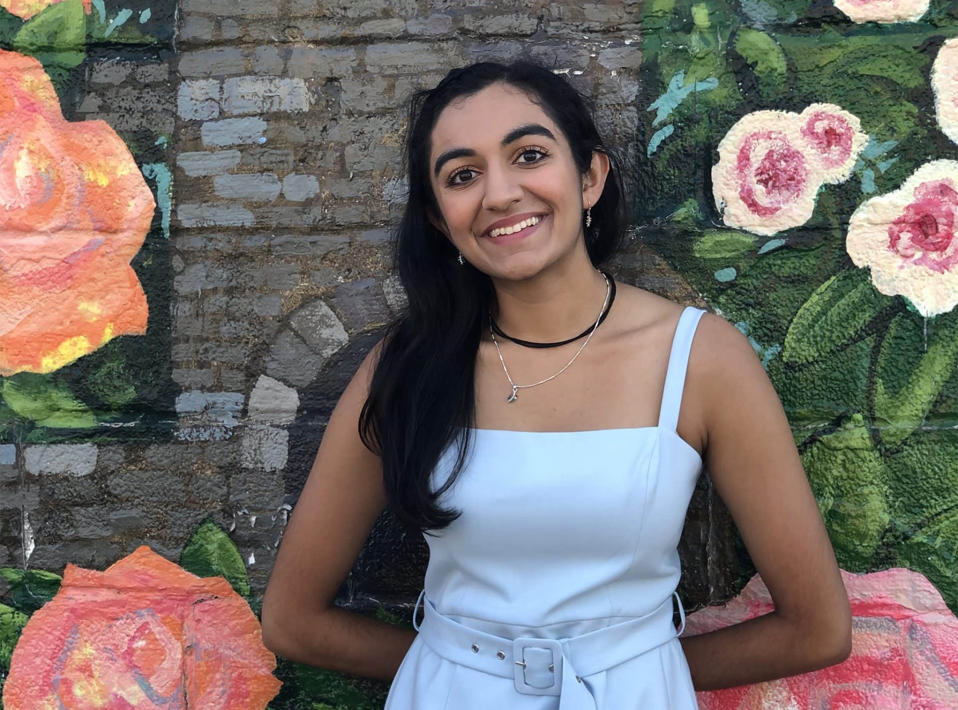 Sanya Shah ’22 smiles for the camera with a mural behind her. The mural has roses and grey brick. She is wearing a light blue dress.