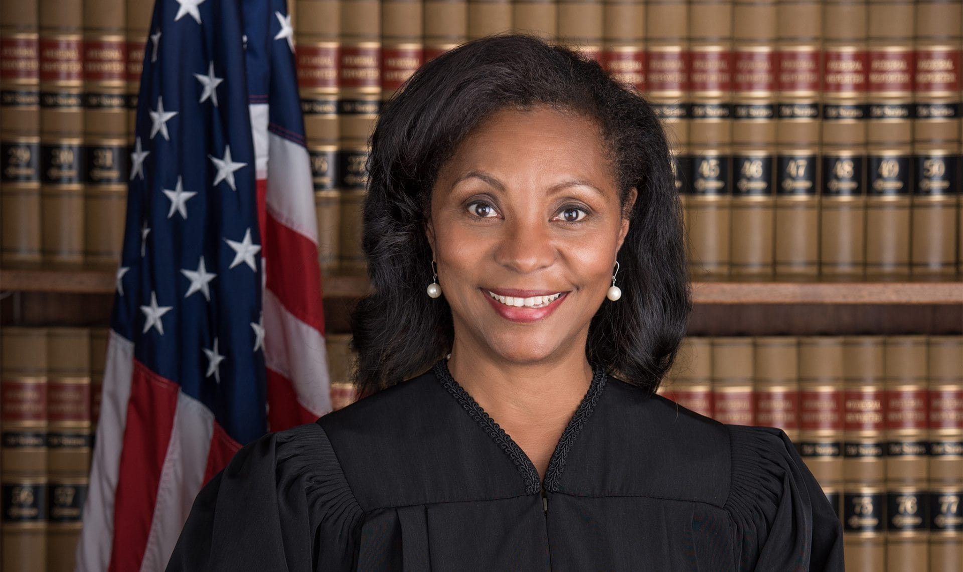Karen Stevenson smiling in a judge's rob in front of rows of legal books and an American flag.