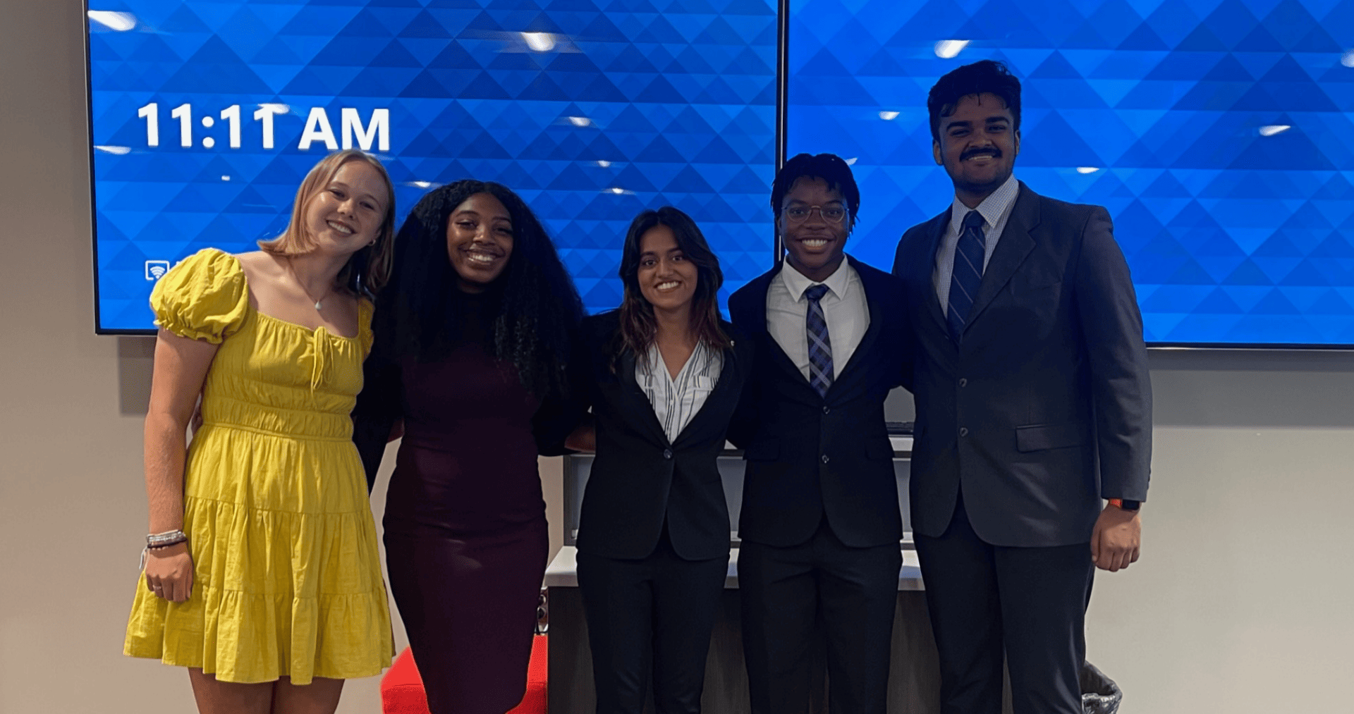 A team of five scholars wearing business attire stand in front of a screen at their final presentation.