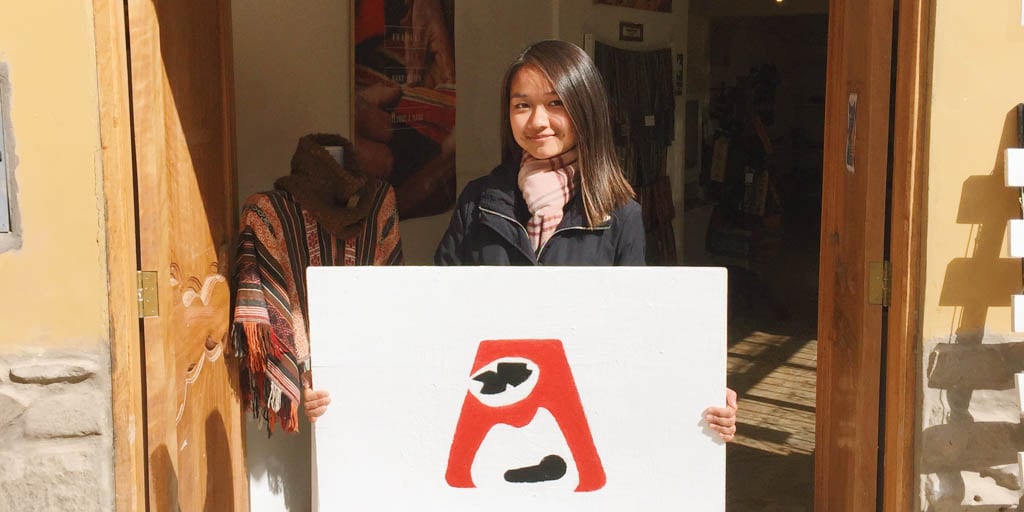 Sara holds a poster-sized sign outside a building. Just inside is a woven poncho on display. There is artwork on the walls.