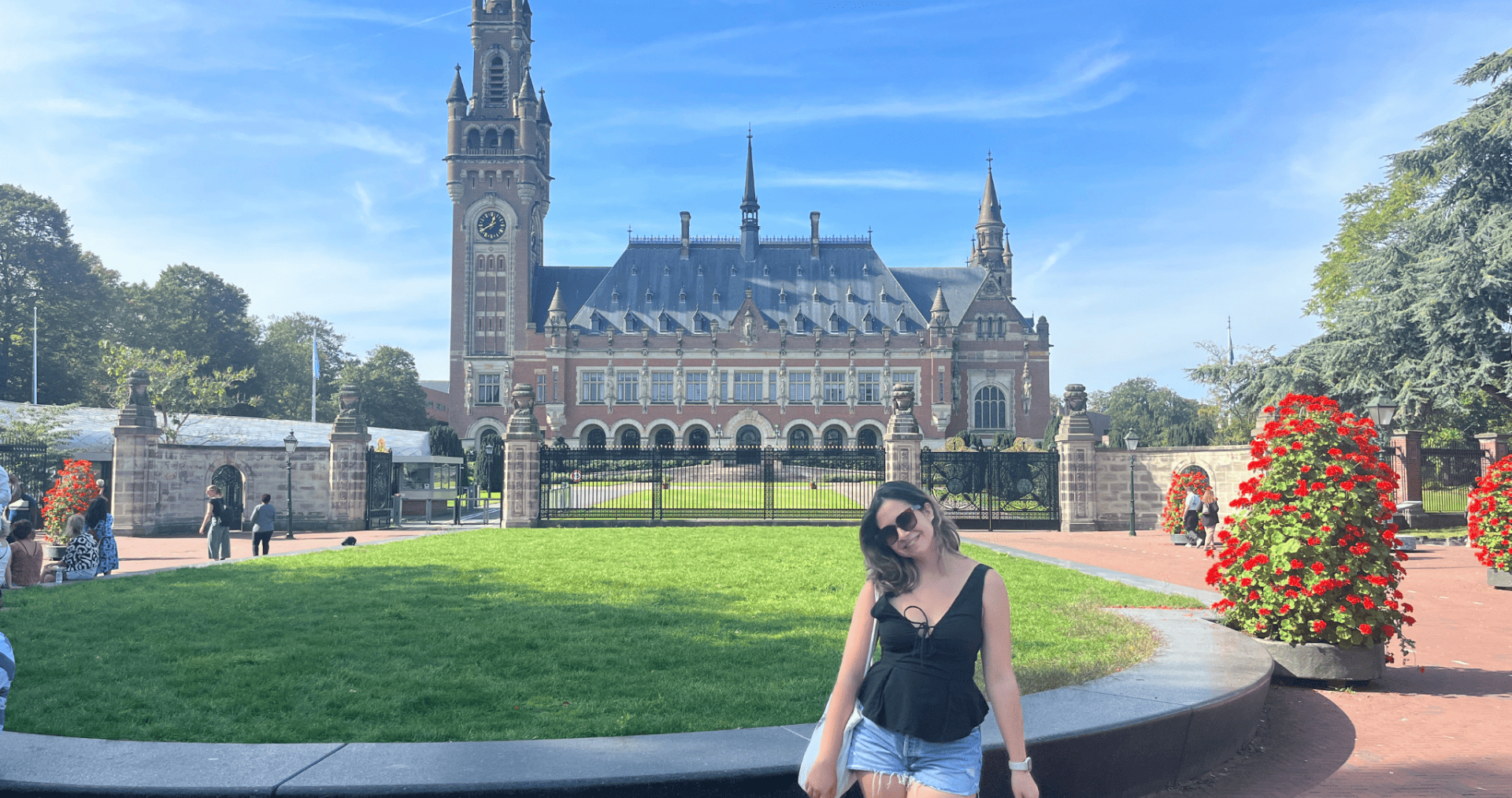 A scholar wearing a black shirt and jean shorts stands outside on a sunny day. She is smiling at the camera. There is a manicured circular lawn, large pots of red flowers, and an ornate building in the background.