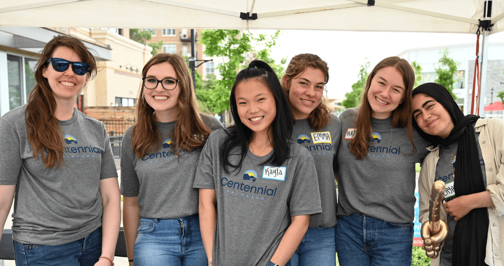 A group of six women wearing matching grey Centennial t-shirts stand and smile for the camera underneath a white tent outside.