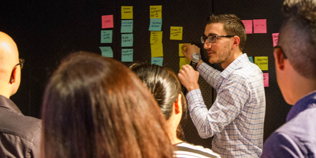 Chris Sopher ’10 points to a Post-it note on the wall in front of a group of people. He is holding a mic and wearing business attire. There are several columns of Post-it notes on the wall.