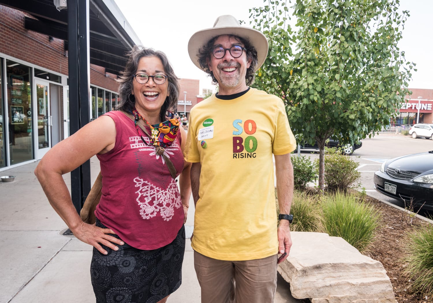 Two smiling people stand on the sidewalk of a shopping center.