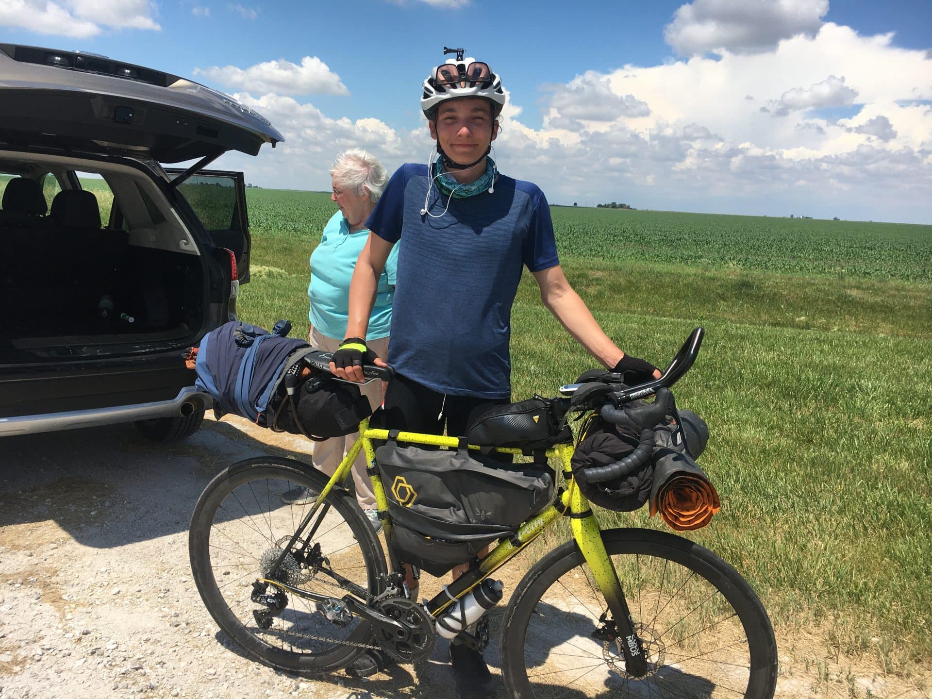 Emmaus smiles while standing behind his bike on a dirt road besides a grassy field.