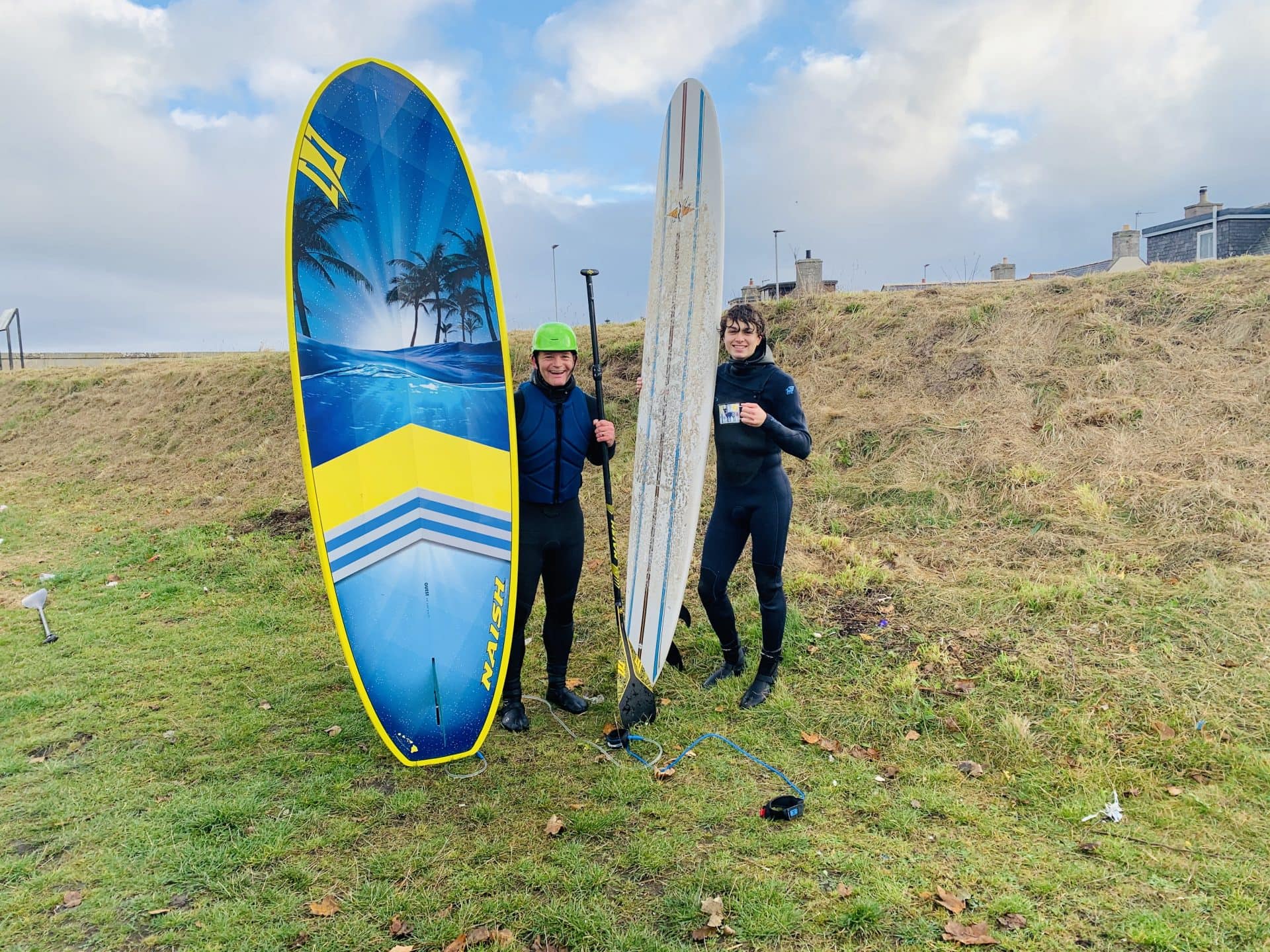 Two people pose for a photo on the coast with surfboards.