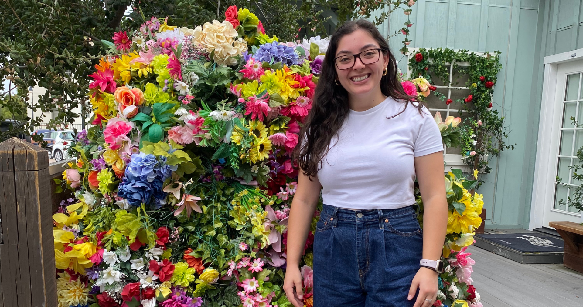 Taylor Molina ’25 smiles for the camera outside with a fixture of brightly colored flowers behind her. She is wearing a white tee and jean shorts.