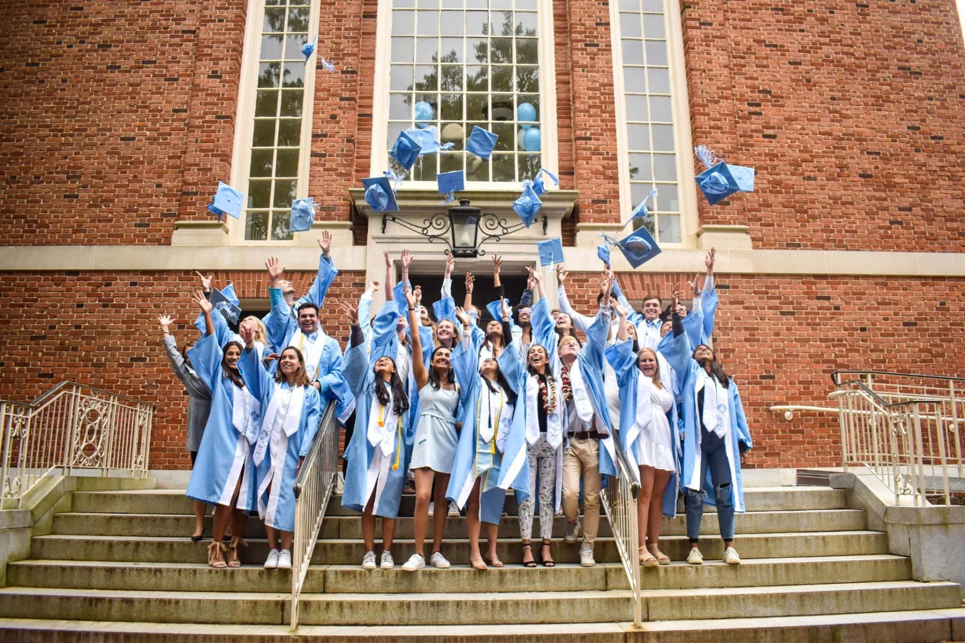 A group of graduates throw their Carolina blue caps into the air in unison on the steps of the Foundation.