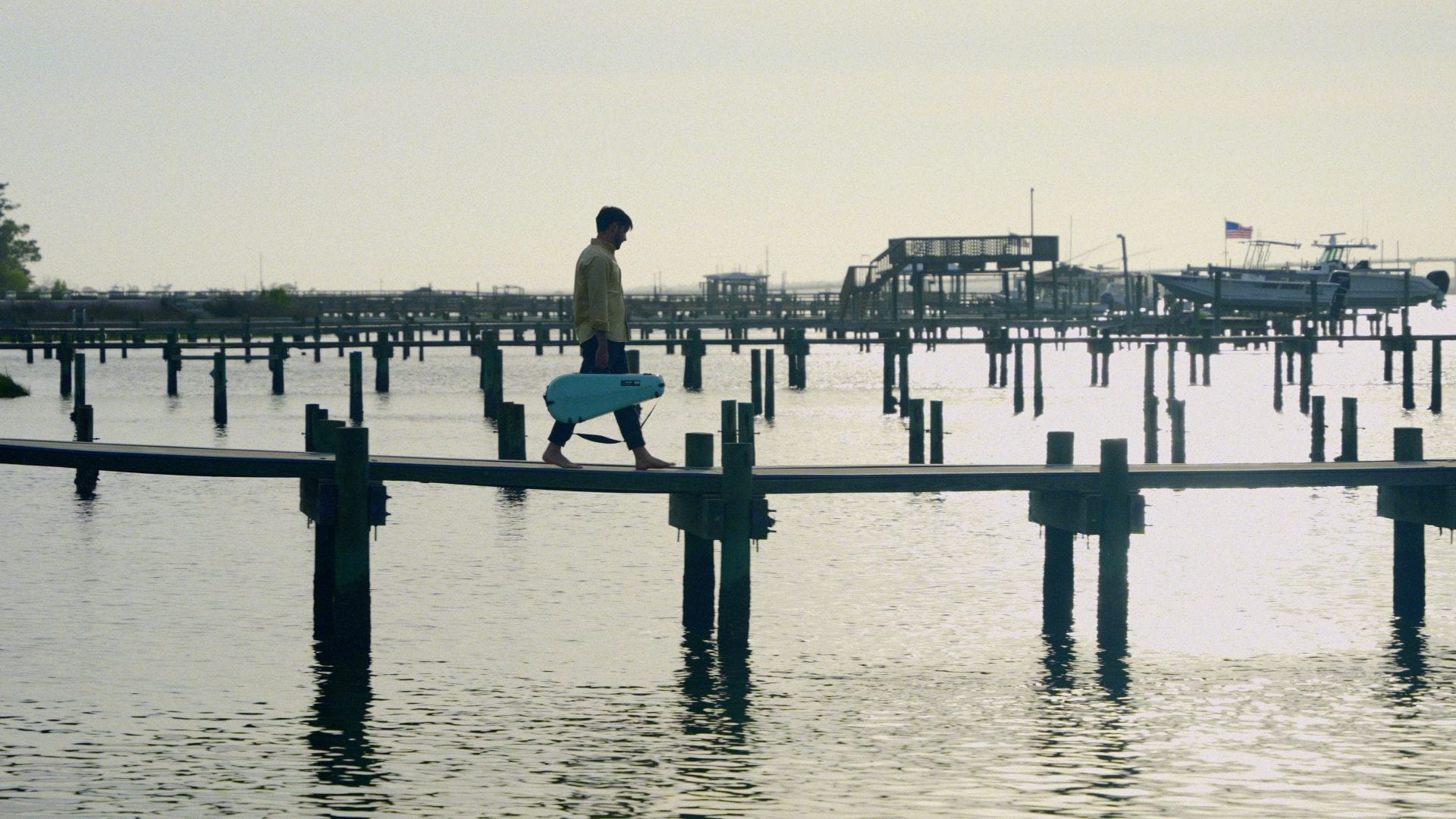 A man carrying a cased mandolin walks across a pier above water at sunset.
