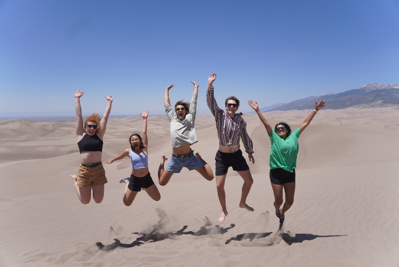 Five scholars jump for a photo on a sand dune.