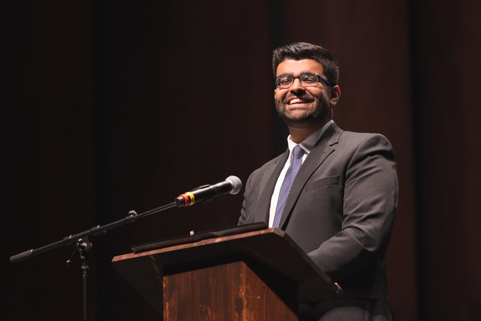 A man dressed in a black blazer, white-collared shirt, and navy tie stands behind a podium with a microphone. He is smiling at the audience.