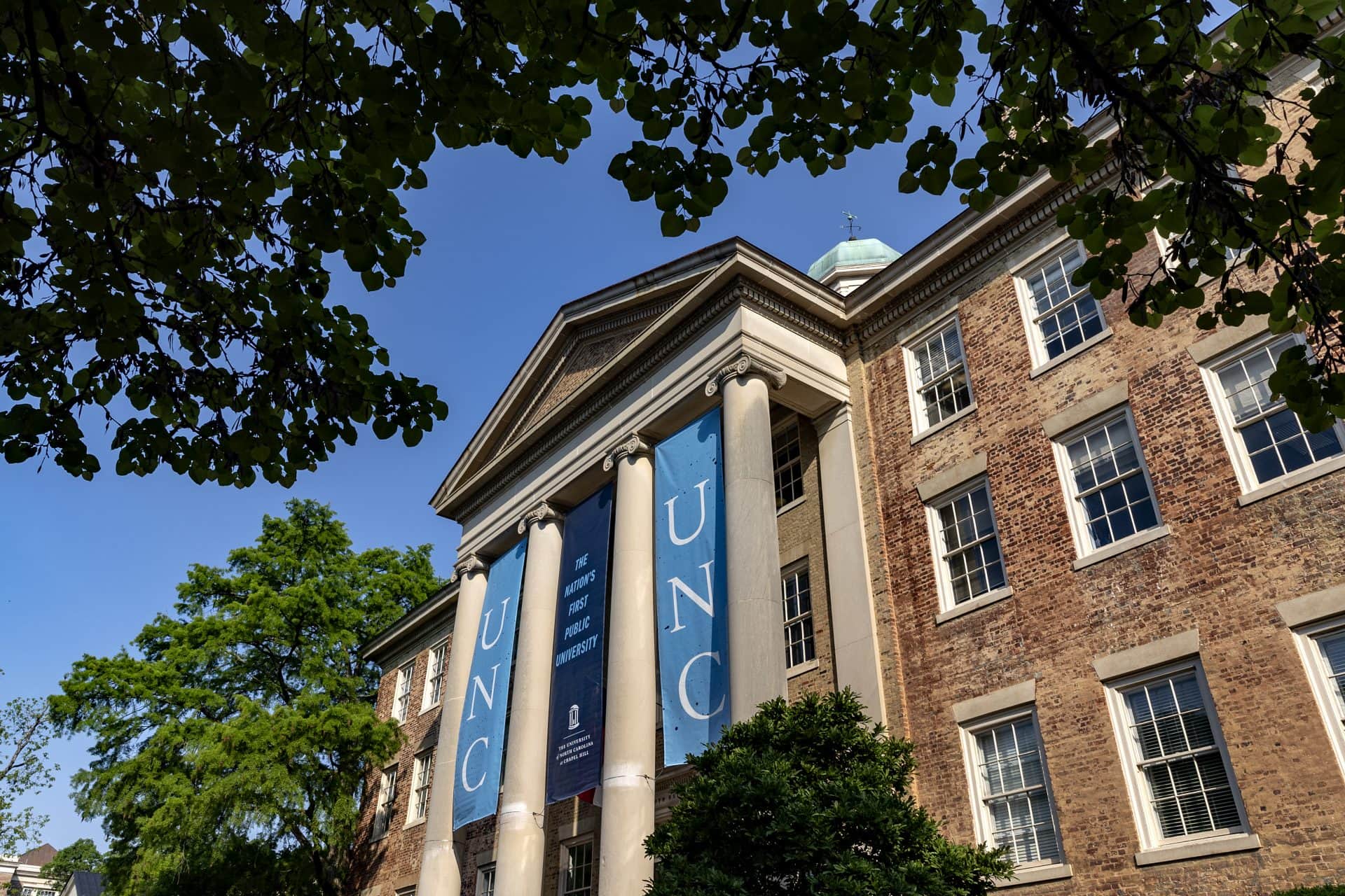 Three UNC banners hang on South Building on the campus of UNC–Chapel Hill. The building is brick with four roman pillars around the main door. Trees surround the building.