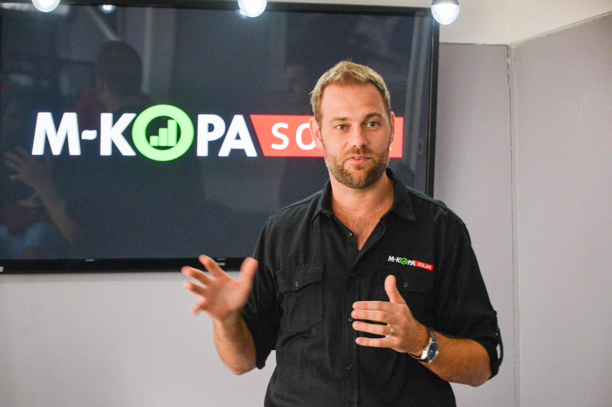 Jesse Moore speaking in front of a television displaying the M-KOPA logo.