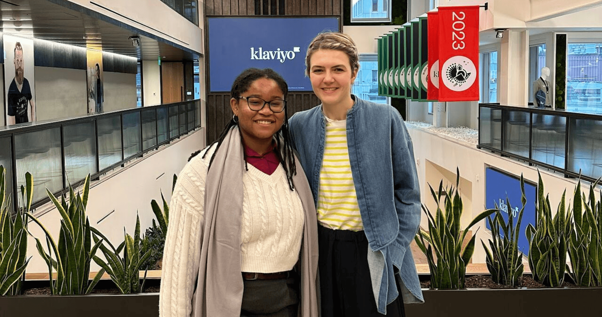 Two women stand inside the Klaviyo offices. There are snake plants and a screen behind them. 