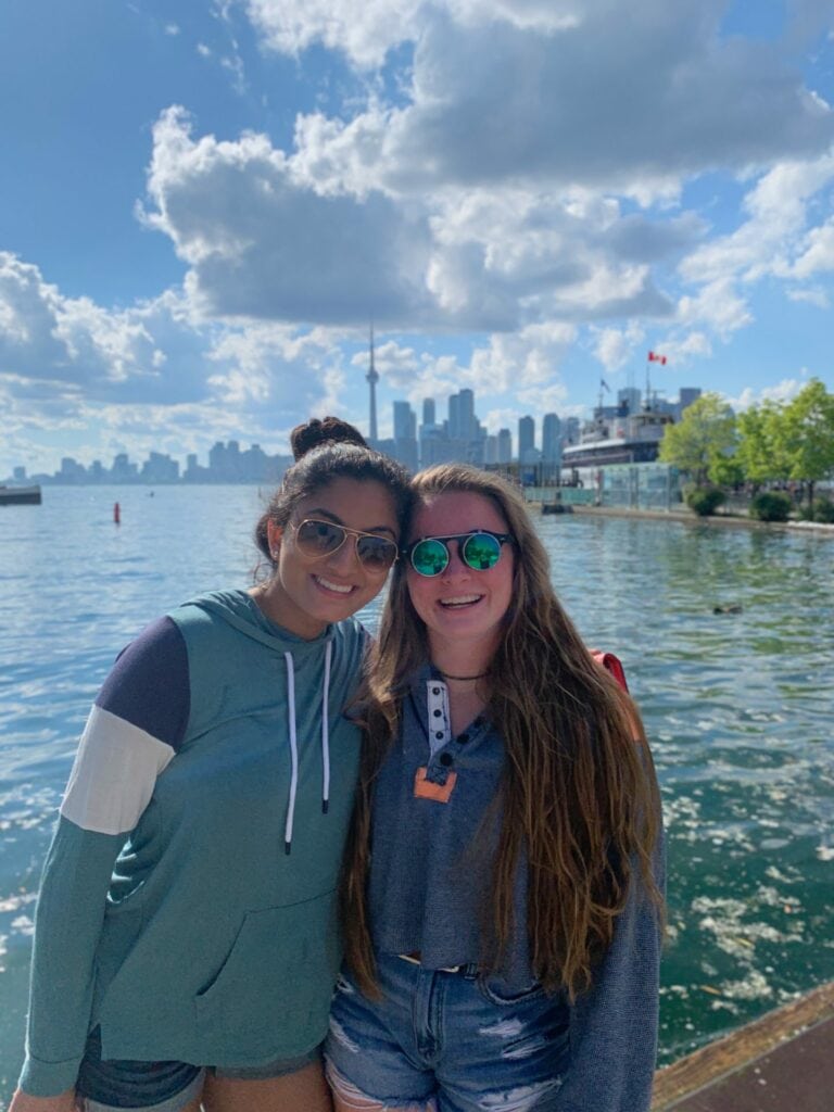 Jaya and Megan smile for the camera by Toronto's waterfront. There are city buildings and trees in the background on a sunny day.