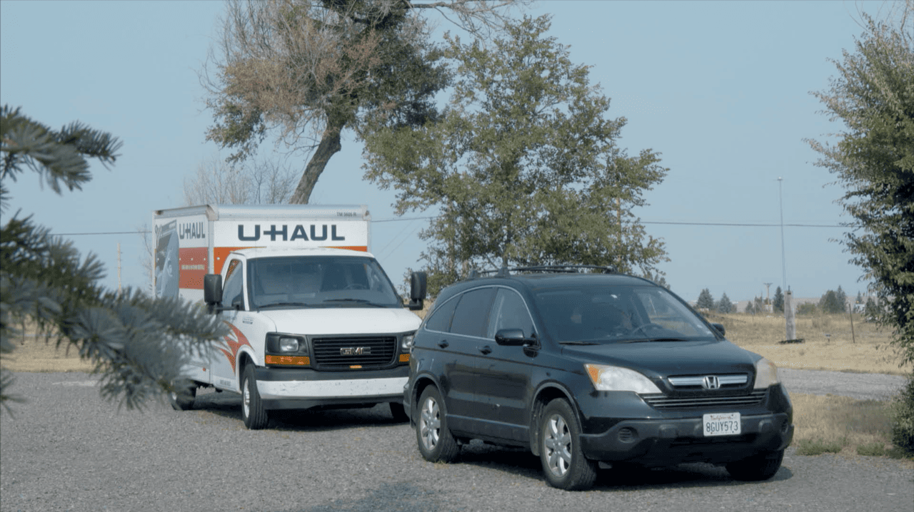 A U-Haul van and a car parked with trees in the background.