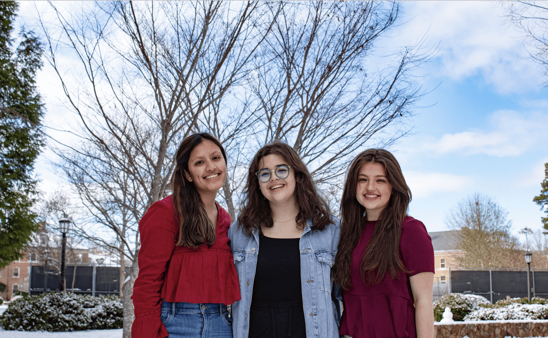 Three women stand outside on a snowy day. There are trees in the background.