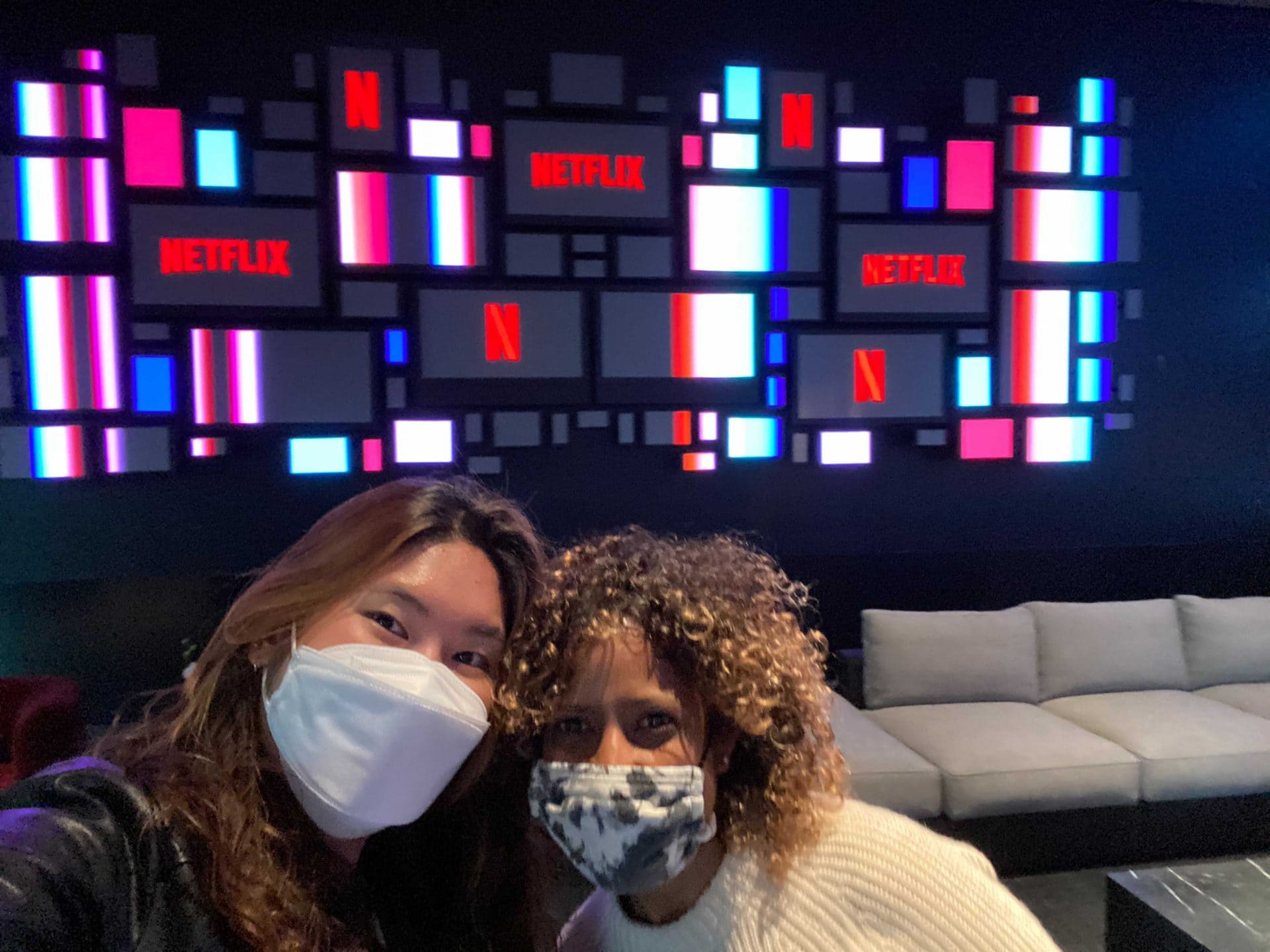 Two women wearing face masks (to protect against transmission of COVID-19) sit inside Netflix headquarters. There is a couch behind them. The wall in the background has lit-up screens with the Netflix logo.