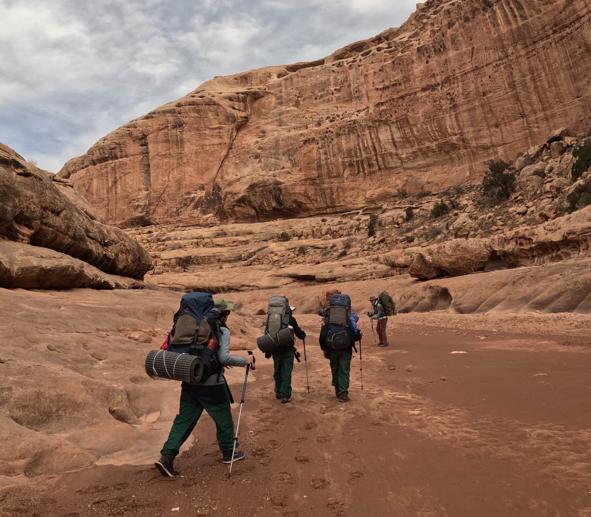 Four backpackers hike through a canyon in the Utah wilderness on a cloudy day. They are carrying hiking sticks, backpacks, and sleeping mats.