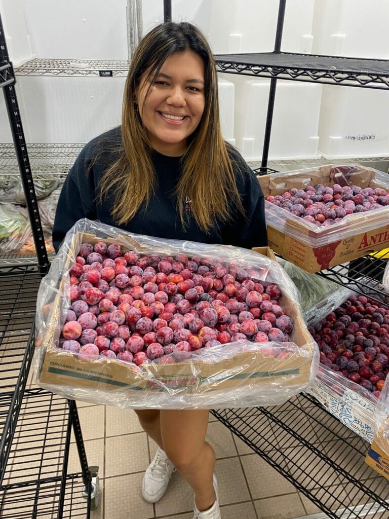 Jenny holds a box of fresh nectarines in a kitchen storage area.