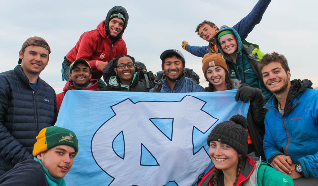 A group of scholars stand outside holding a large UNC flag. 