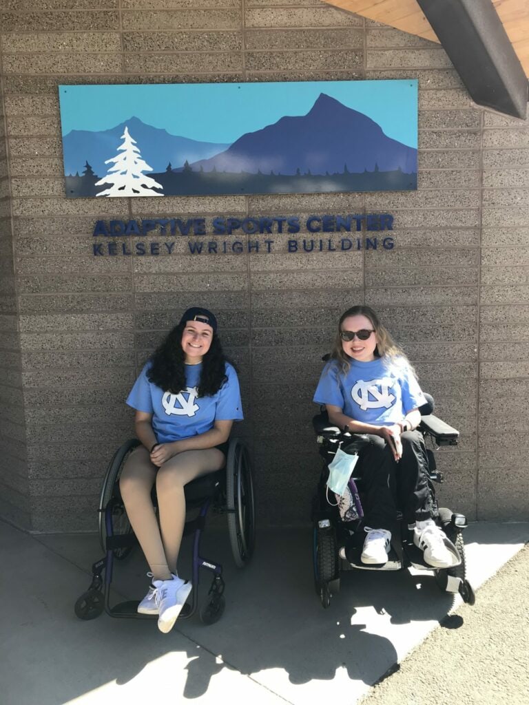 Laura and Eleanor smile in front of the Adaptive Sports Center building wearing Carolina t-shirts.