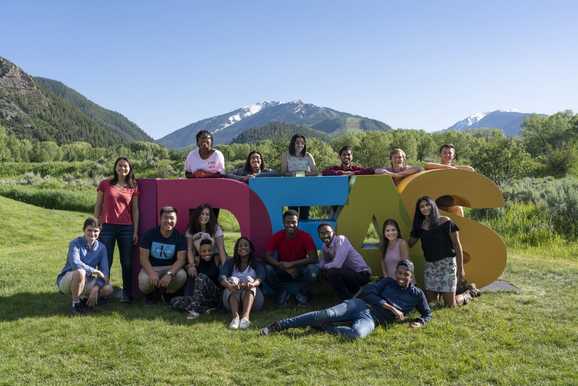 A group of students surround an installation (four letters that read ”D-E-A-S”) on the grass. There are mountains and trees behind them. All of are smiling.
