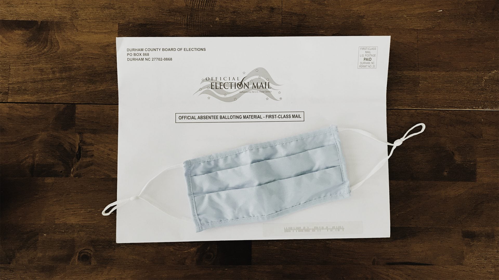An absentee ballot envelop sits on a wooden table with a cloth face mask on top