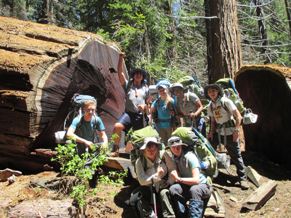 A group of students wearing hiking gear smile at the camera in the forest.