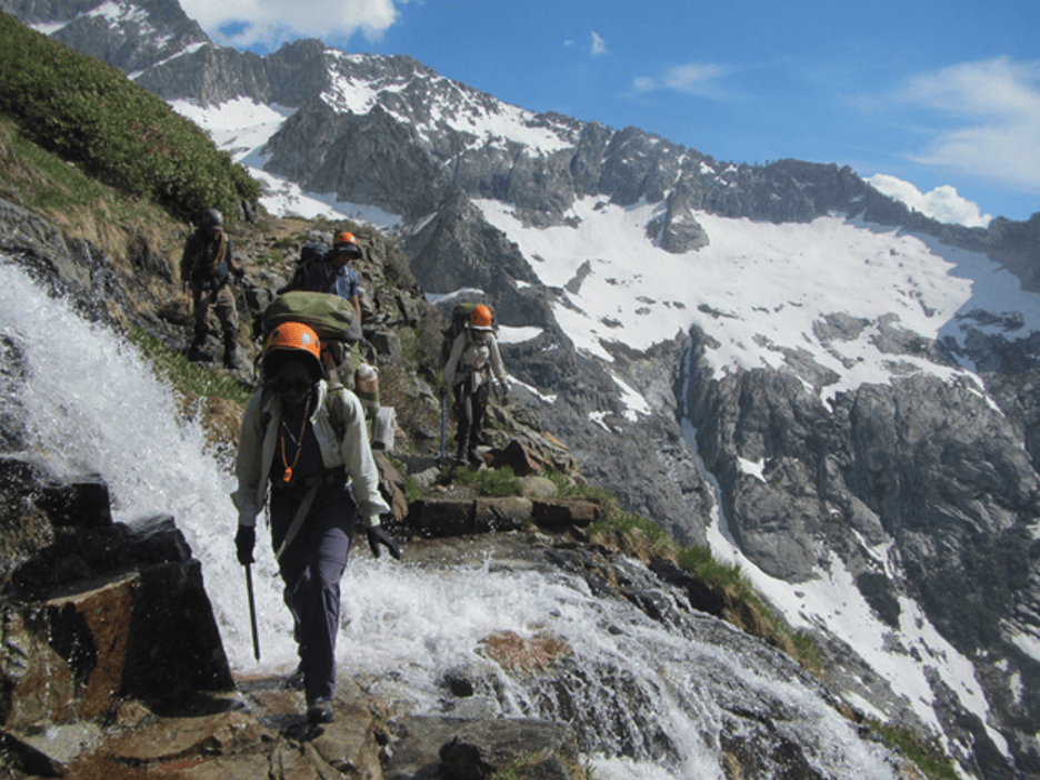 Three hikers clamber over the side of a rocky and snowy mountainside.