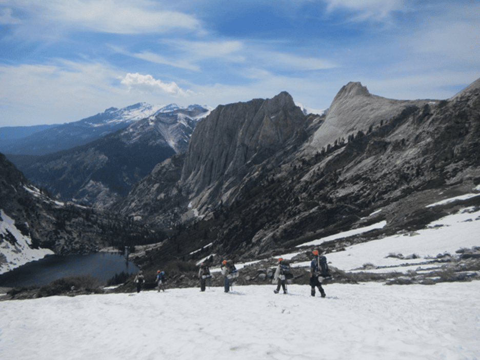 Backpackers walk through snowy mountains towards a lake. 