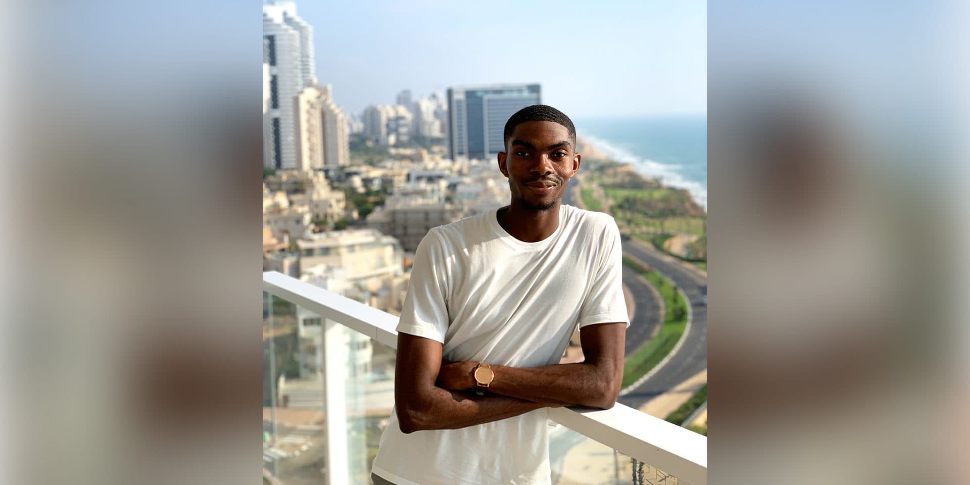 Uzorma Owete ’20 smiles softly for the camera with a city view behind him. The coast is also in view next to the road. He is standing on a balcony on a sunny day.