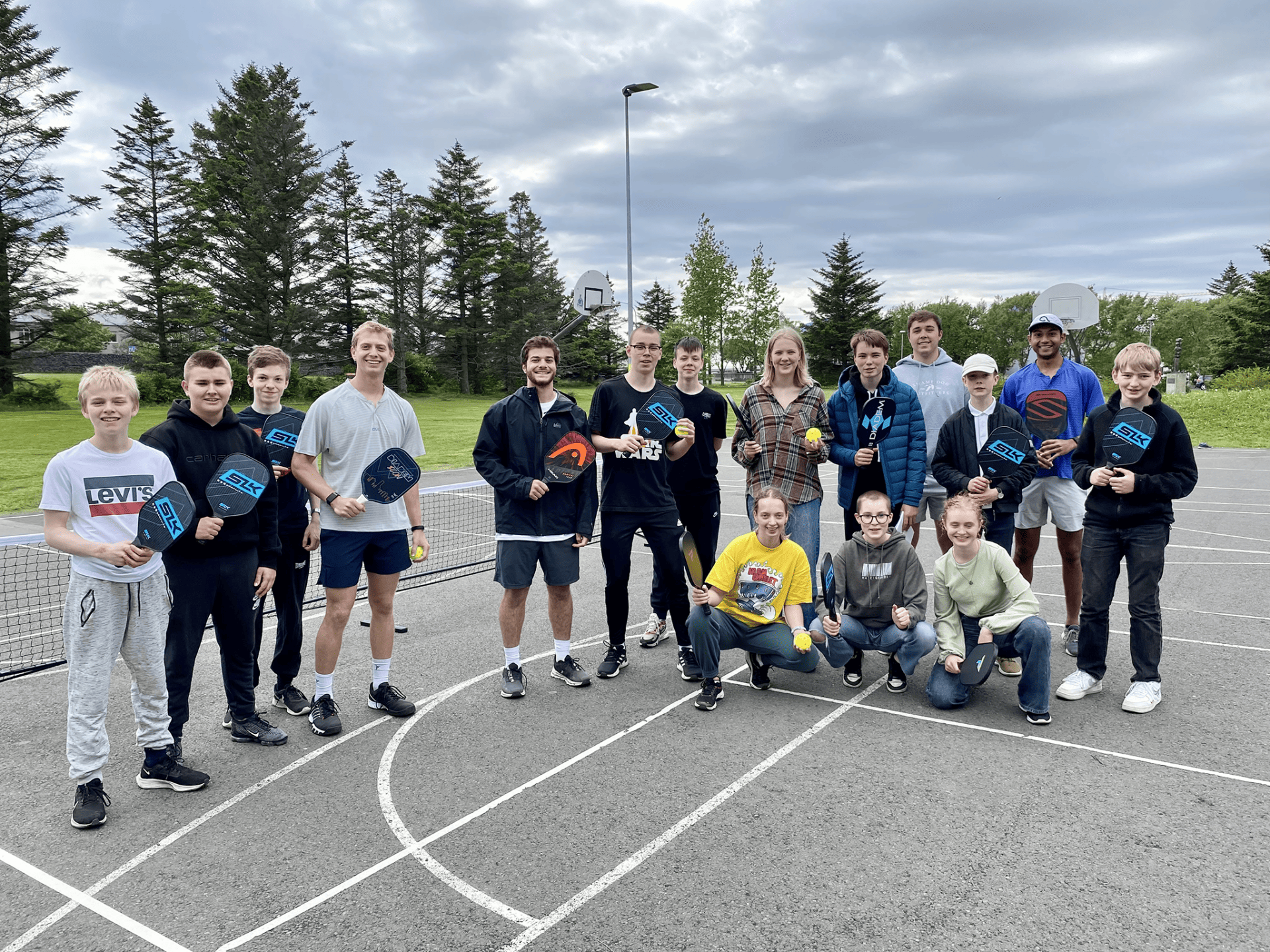 A group of pickle ball players smile for the camera with their paddles. They are standing on a tennis court on a cloudy day. There are trees behind them.