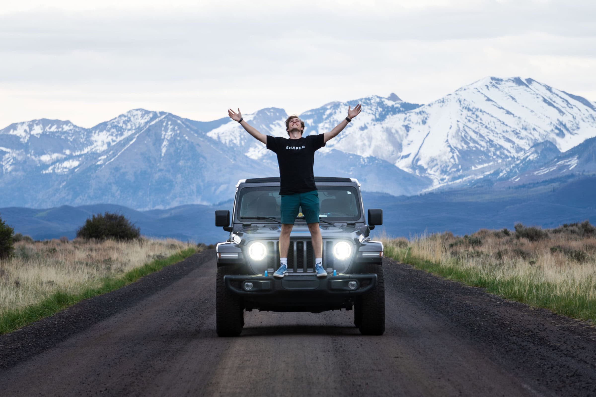 Jake stands on his Jeep on a road with snowy mountains behind him. His arms are stretched out wide, and he’s smiling.