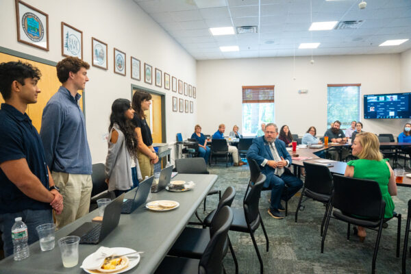 Four college students stand before the Western North Carolina regional council board meeting, a large group of adults seated at tables