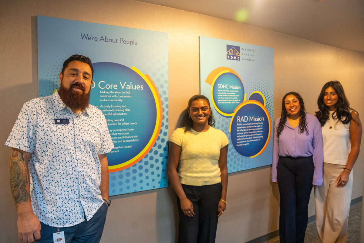 An adult male and three female college students smile with two infographic poster boards about the San Diego Housing Commission's mission and values