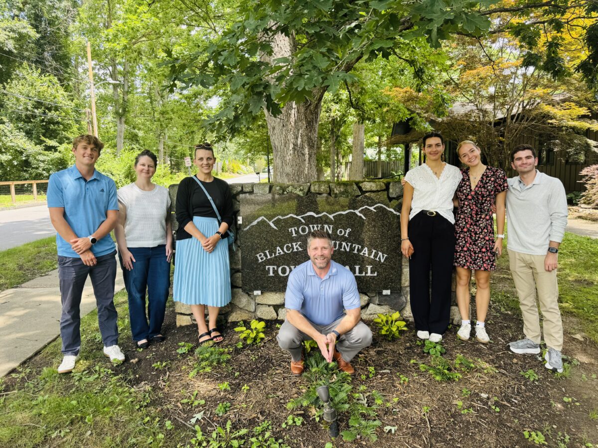Four college students and three adults smile with the Town of Black Mountain Town Hall sign