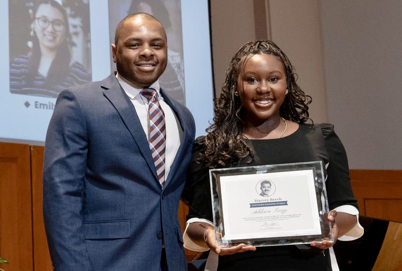 A man and woman in business attire stand on stage. The woman is holding her Harvey Beech Scholarship certificate.
