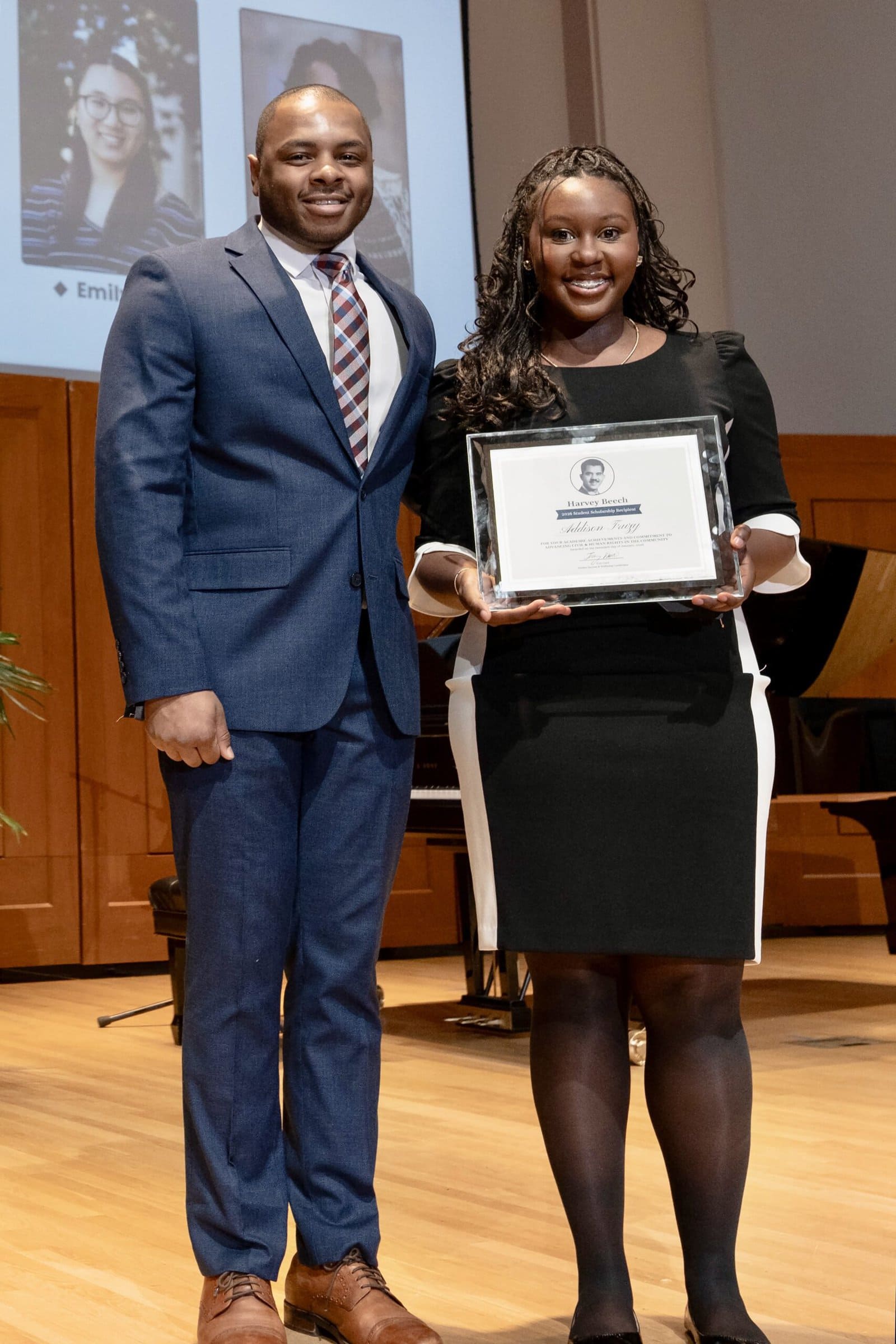 A man and woman in business attire stand on stage. The woman is holding her Harvey Beech Scholarship certificate.