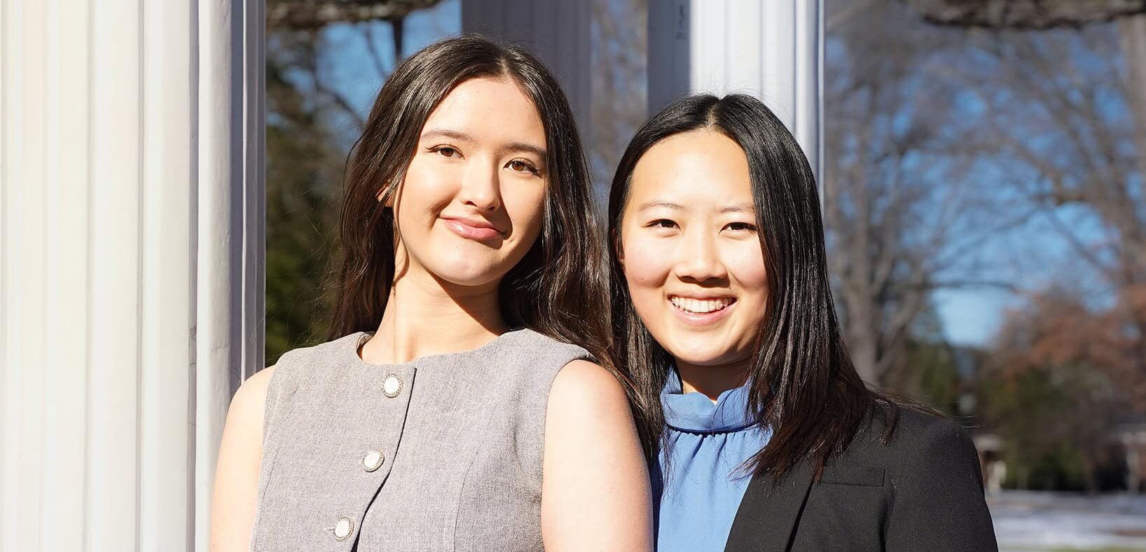Two female college students stand in front of the Old Well wearing business attire.