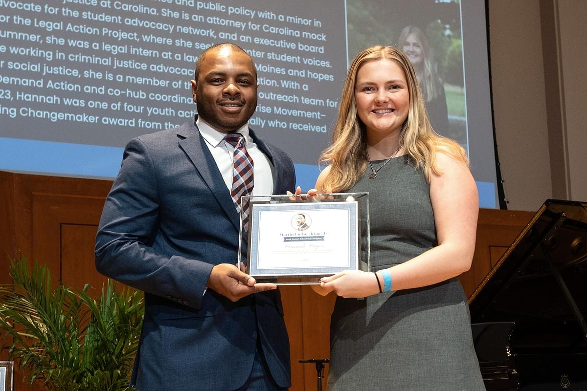 A man and woman in business attire stand on stage. The woman is holding her MLK UNC Student Scholarship certificate.