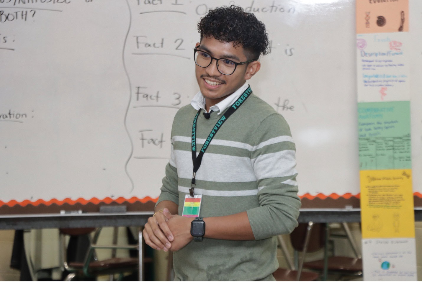 A teacher smiles at students with a whiteboard behind him.