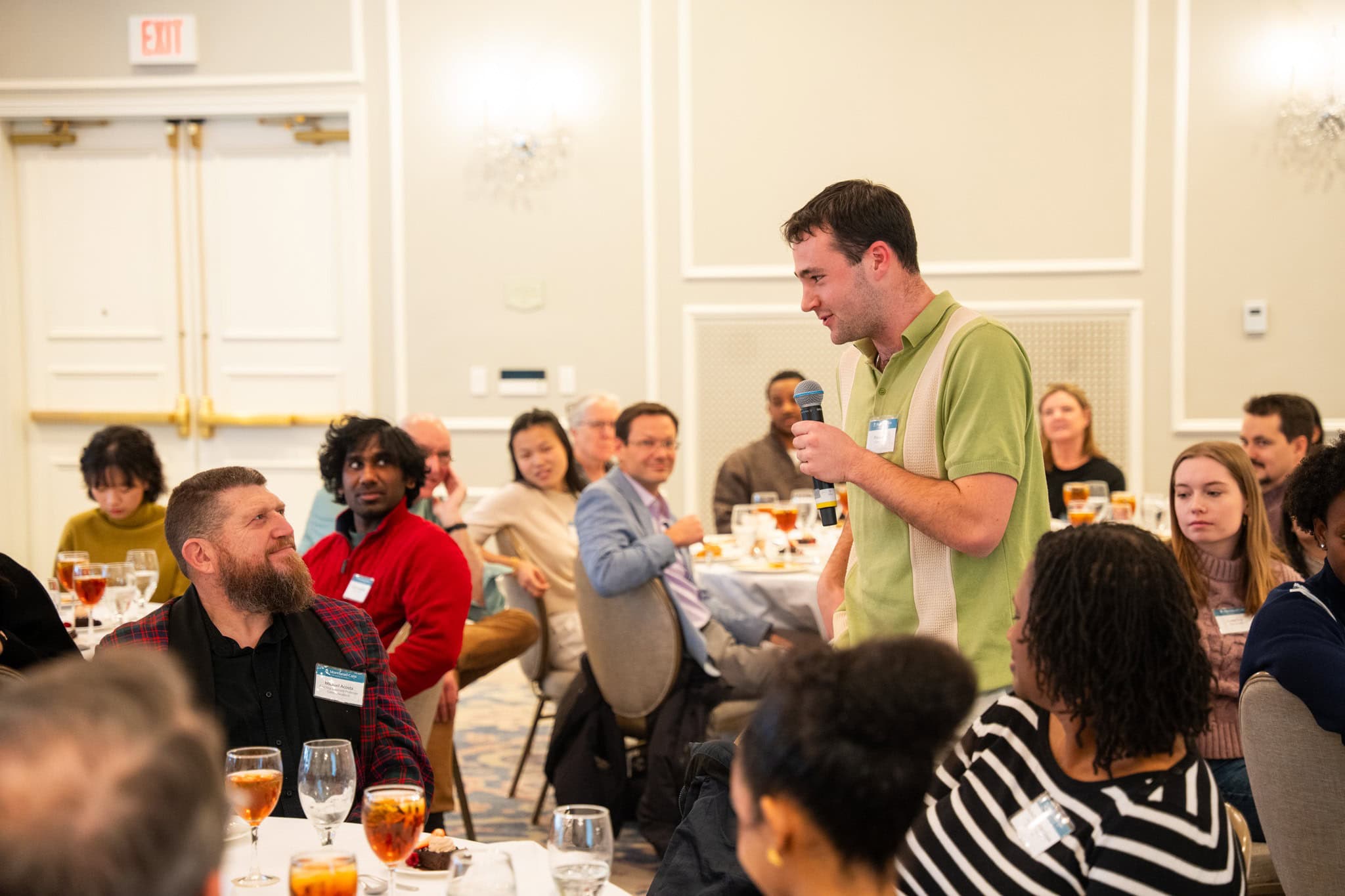 Finn Sansom stands holding a microphone at a luncheon at the Carolina Inn, sharing remarks as his professor, Michael Acosta, looks on with a smile. Attendees seated at tables watch in the background.